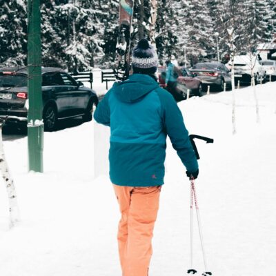A person in winter clothing carrying skis and poles on a snowy street.