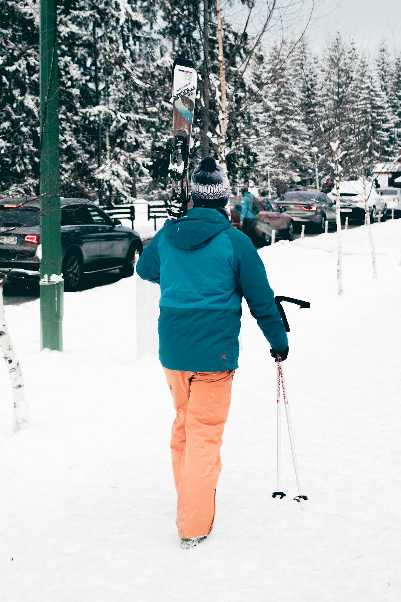 A person in winter clothing carrying skis and poles on a snowy street.
